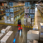 Aerial view of a warehouse with workers in safety vests and helmets conversing. Forklifts and stacked boxes fill the space, symbolizing logistics and industrial storage. The organized layout highlights efficient distribution operations.